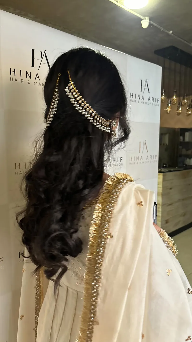 Back view of long, loose curls finished with pearl and gold hair chains, in front of the salon’s Hina Arif monogram backdrop